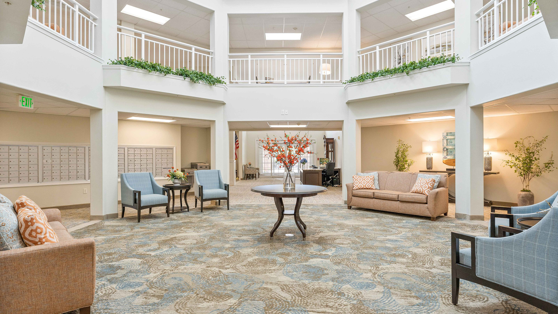 Bright, spacious lobby with a round table and floral arrangement in the center, surrounded by sofas and chairs. Upper level with white railings, plants, and natural light from windows above.