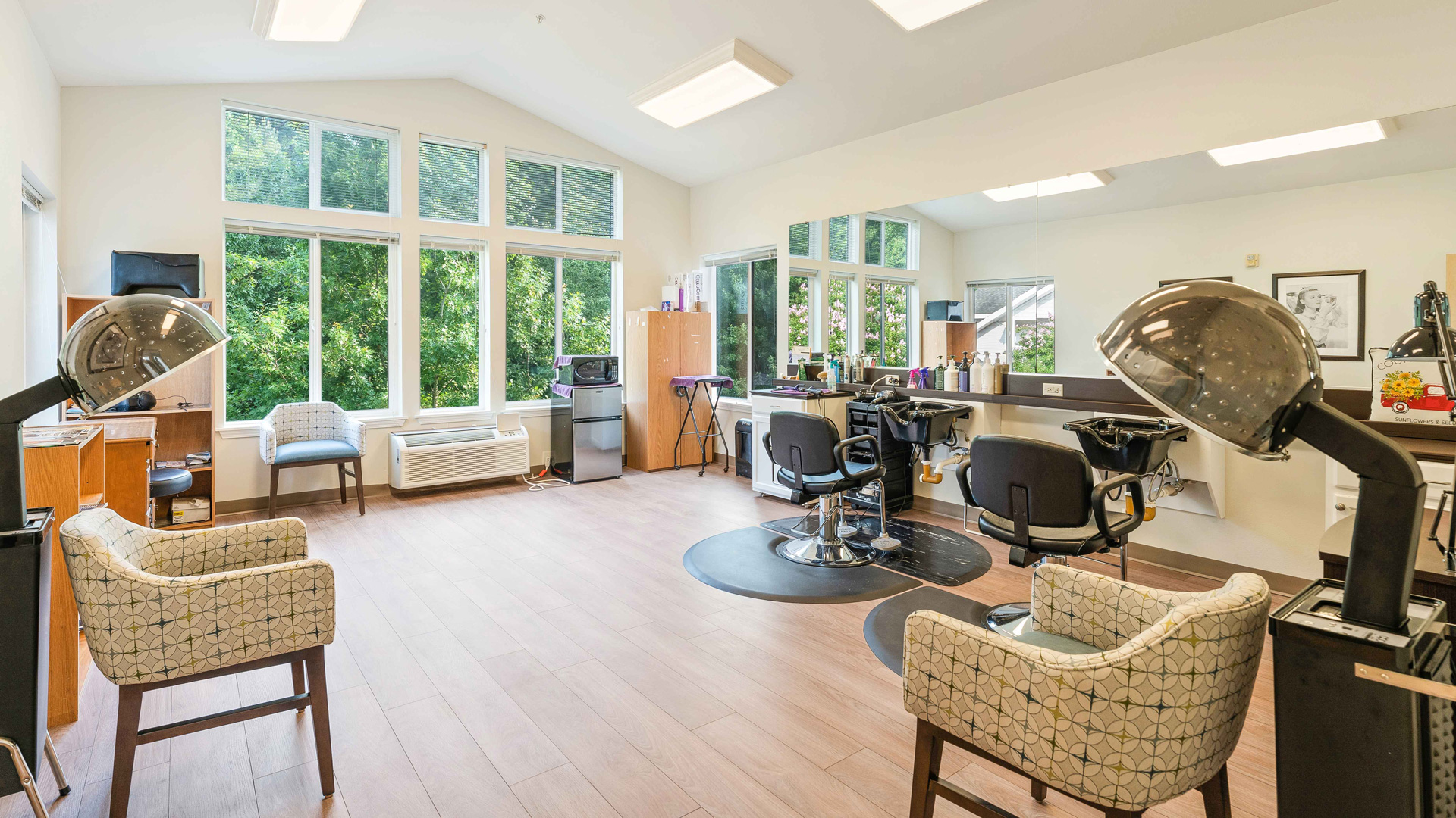 Bright, modern salon with large windows, two styling chairs with mirrors, two vintage hair dryers with chairs, and wooden flooring. Shelves, a mini fridge, and greenery visible outside add to the inviting atmosphere.