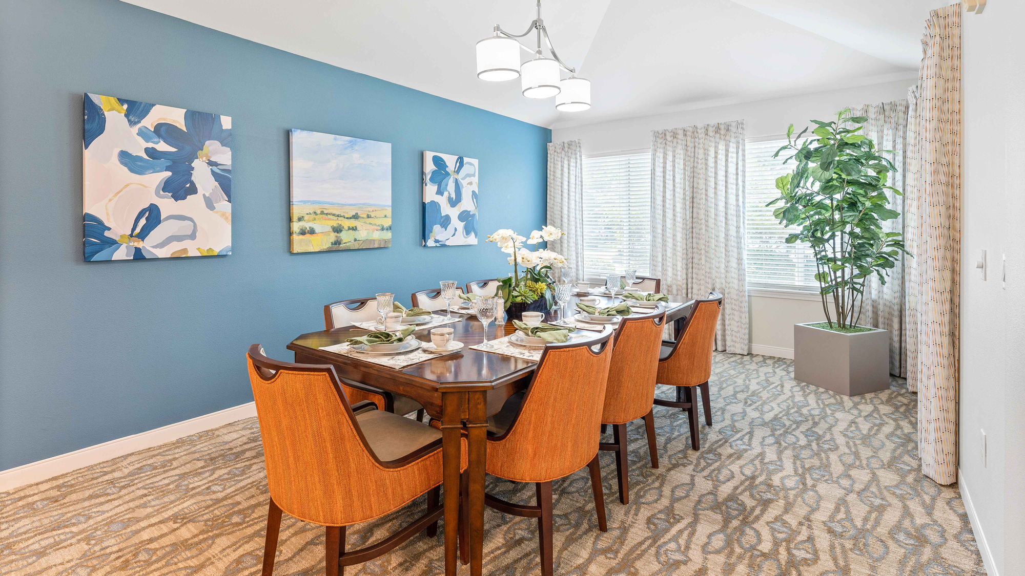 A modern dining room with a blue accent wall, three floral paintings, a rectangular wooden table set for eight, orange chairs, patterned curtains, a large potted plant, and bright natural light from windows.