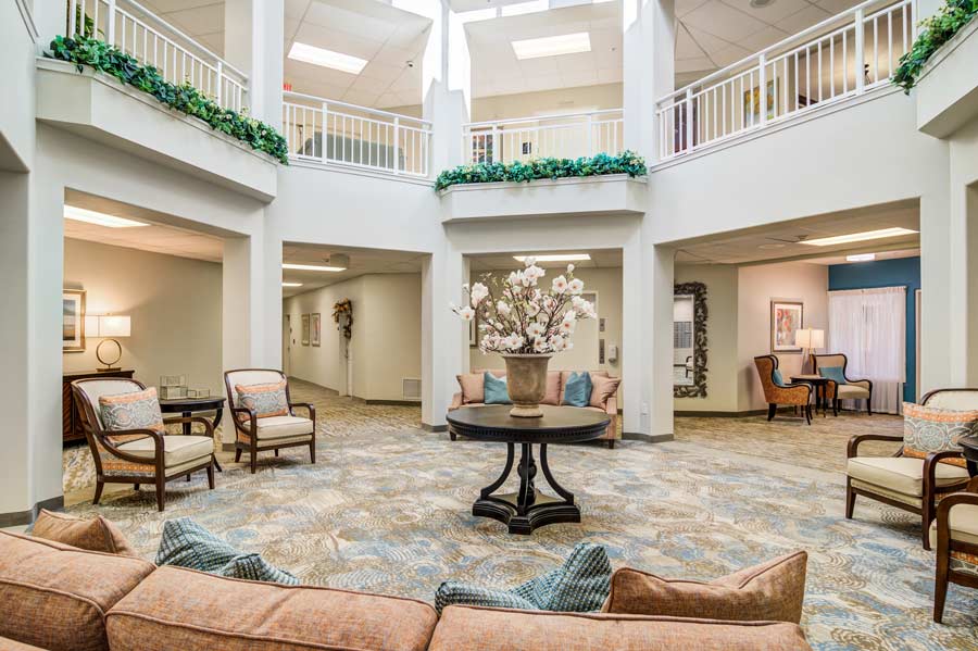 A spacious, well-lit lobby with a high ceiling, decorated with potted plants and cozy seating areas. The central round table displays a vase filled with white flowers. The walls and floor are painted in neutral tones, giving a calm and inviting atmosphere.