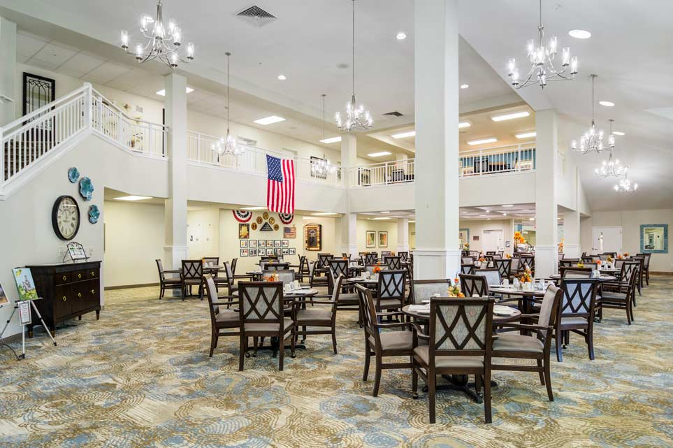 A spacious, well-lit dining area with numerous tables and chairs. Chandeliers hang from the high ceiling, and an American flag is displayed prominently on a second-floor balcony. The decor is elegant, featuring framed pictures and various other decorations.