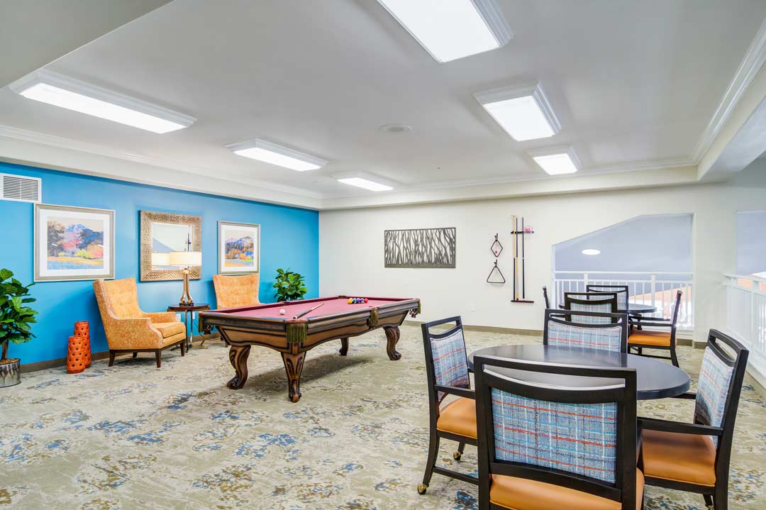 A recreational room with a pool table in the center, surrounded by chairs and small tables. The walls are decorated with framed artwork, and the room is well-lit with ceiling lights. There are also potted plants and cues hung on the wall.