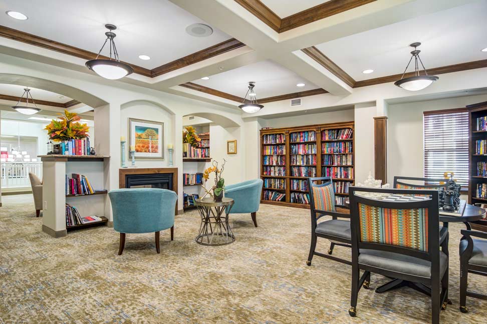 A cozy, well-lit library with modern ceiling lights. It features rows of bookshelves filled with books, a fireplace with decorative pieces, and comfortable seating with blue upholstered chairs and a small table. A window allows natural light to brighten the room.