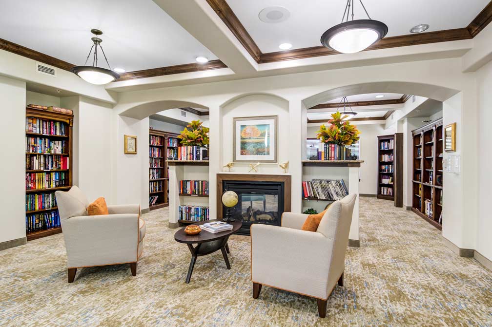 A cozy library with two beige armchairs facing a fireplace. The room features built-in bookshelves filled with books, warm lighting from ceiling fixtures, and autumnal floral arrangements. There is a small coffee table between the chairs on a patterned carpet.