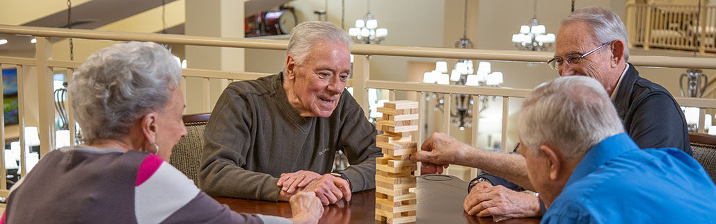 A group of four elderly people are gathered around a table, playing a game of Jenga. They are smiling and look engaged in the activity. The background features a well-lit indoor setting with chandeliers and a beige railing.