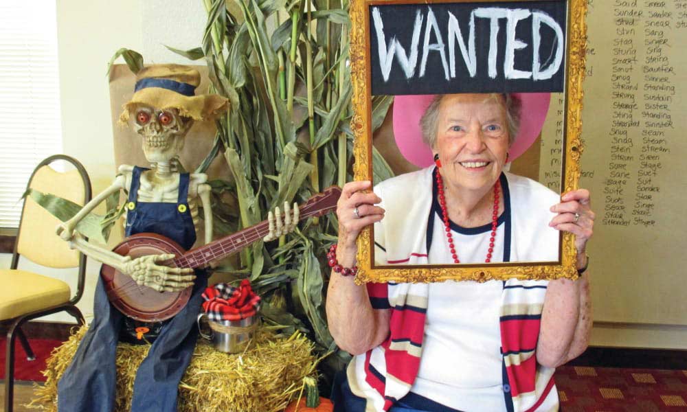 An elderly woman is seated, smiling, and holding a decorative frame labeled 