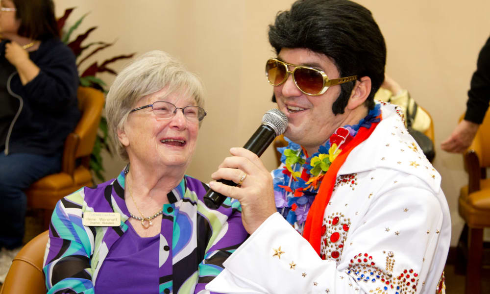 An elderly woman wearing glasses and a colorful top smiles while next to a man dressed as Elvis Presley, who is holding a microphone and wearing a white jumpsuit with ornate details, sunglasses, and a floral leis. People in the background sit on chairs.