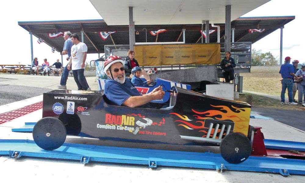 A smiling man with a gray beard and glasses sits in a small race car adorned with flames and sponsor logos. He is wearing a helmet and holding his thumbs up. Other people and spectators can be seen in the background at an outdoor event.