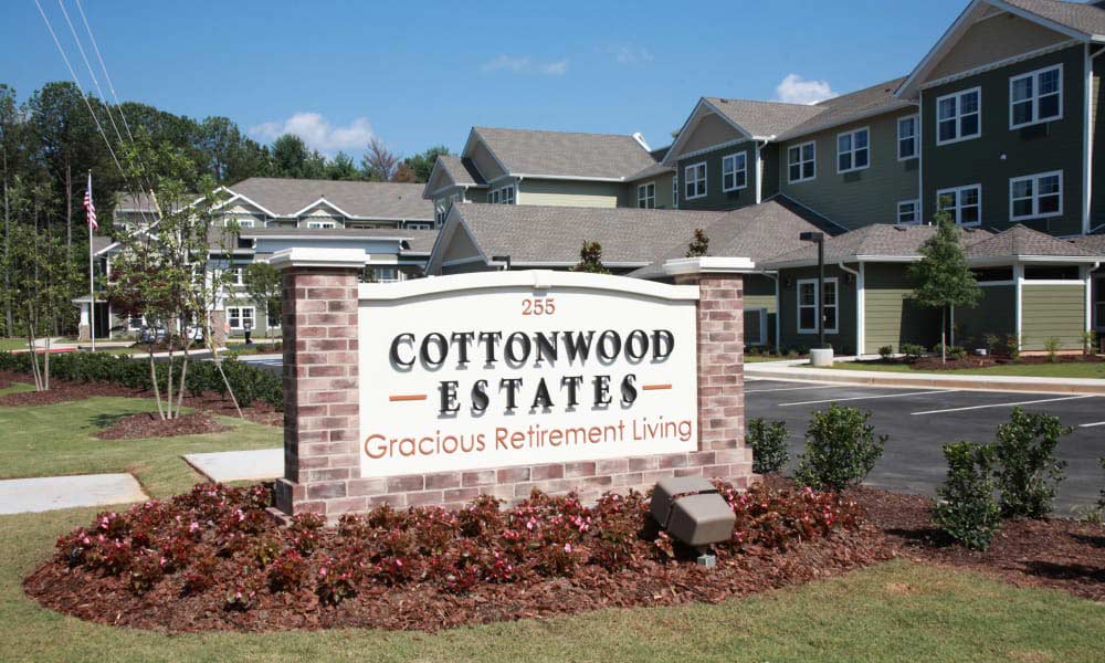 A sign for Cottonwood Estates Gracious Retirement Living is displayed in front of a residential building with multiple floors. The sign is made of brick and white stone, with neatly landscaped bushes and grass surrounding it. The address 