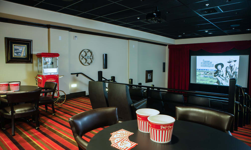 A private home theater room features a large screen displaying a Western movie. Tables with chairs are positioned in front of the screen, each table holding popcorn buckets and red-striped popcorn bags. A popcorn machine is situated against the wall under a framed picture.