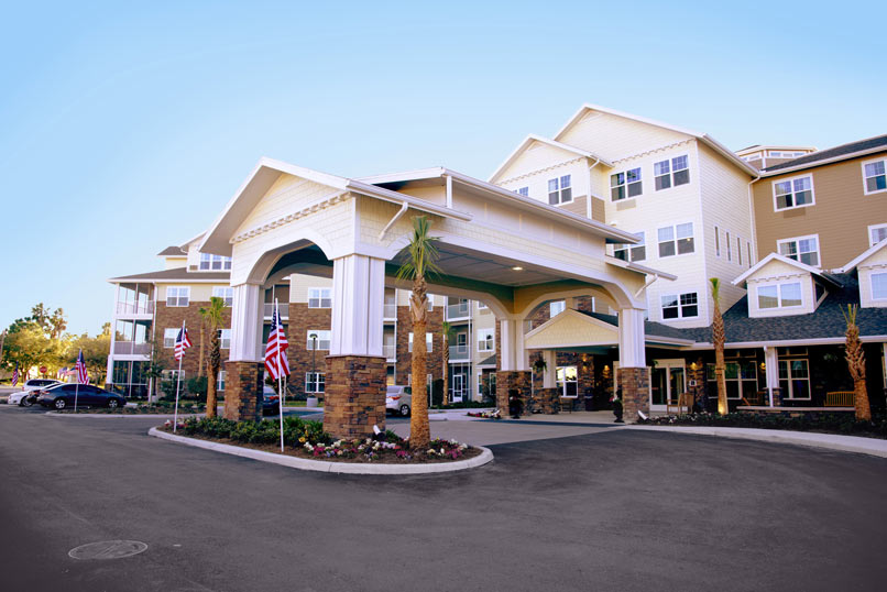 A modern multi-story building with a large covered entrance and an American flag. The structure features beige and white siding and numerous windows. Palm trees and small flower beds adorn the entrance area, and a clear blue sky is in the background.