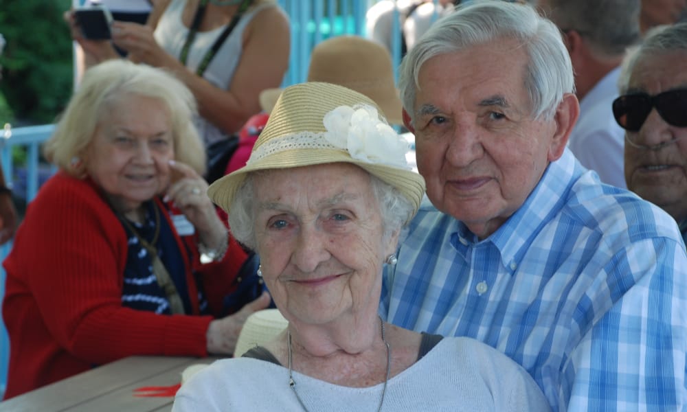 An elderly couple smiling at the camera. The woman wears a hat with a flower, and the man has gray hair and wears a blue plaid shirt. They are seated outdoors at a table, with other elderly people in the background. The atmosphere appears to be cheerful and social.