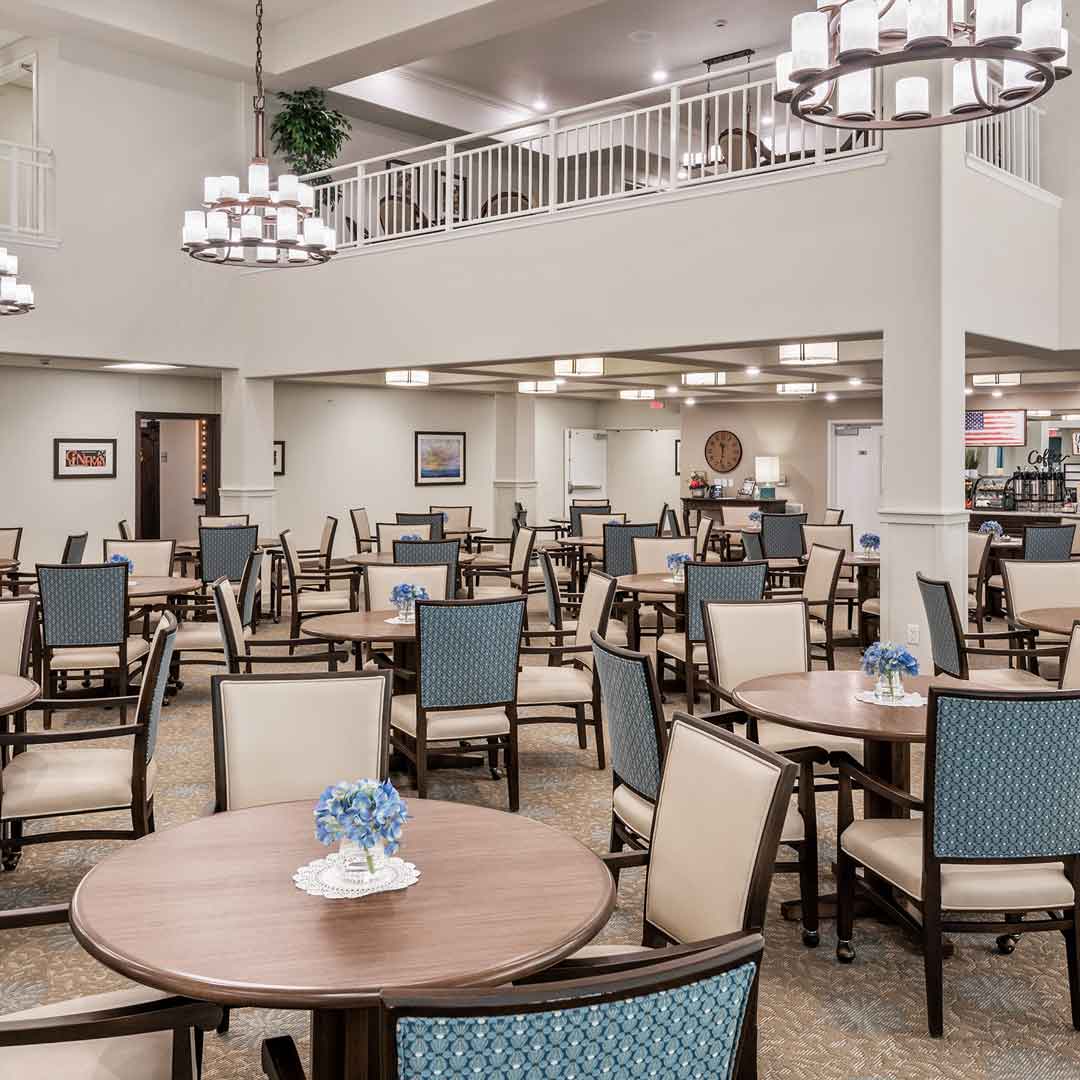 Spacious dining area with multiple tables and chairs, featuring blue flower centerpieces. The room has beige walls, a high ceiling, large chandeliers, and a second-floor balcony. The decor is elegant and inviting.