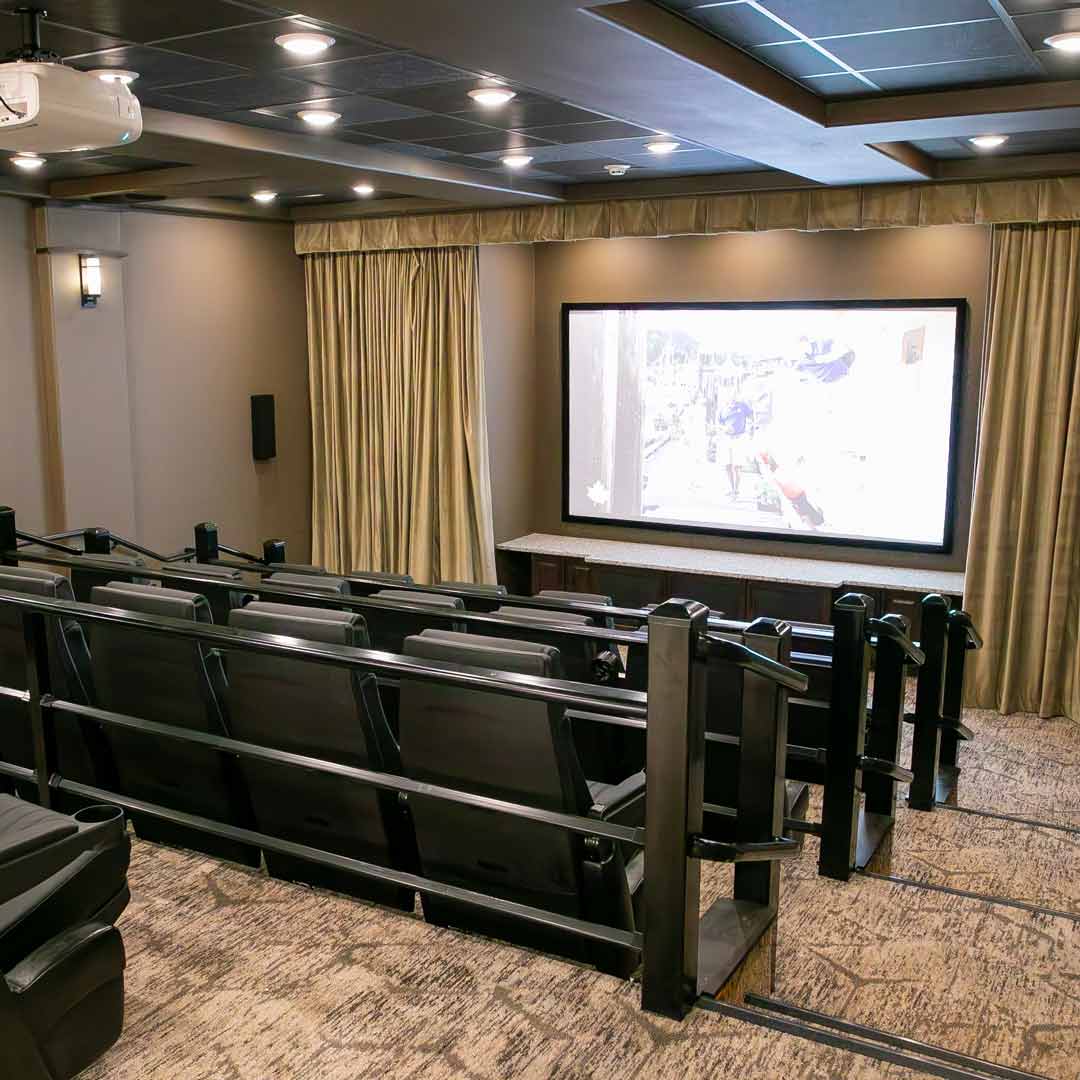 A home theater room with three rows of black leather recliners facing a large projection screen. The walls are painted in neutral tones, and there are beige curtains on the sides. Ceiling lights illuminate the space.