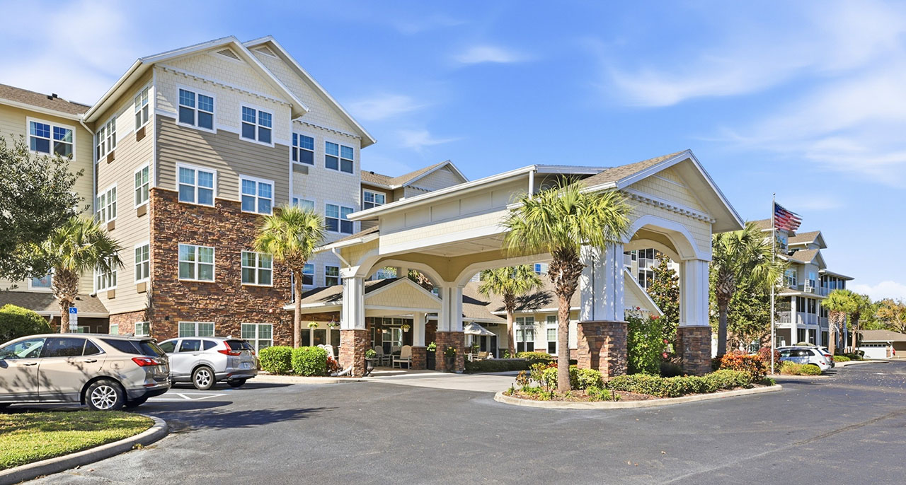 A modern multi-story residential building with a covered driveway entrance, surrounded by palm trees, plants, parked cars, and an American flag on a sunny day.