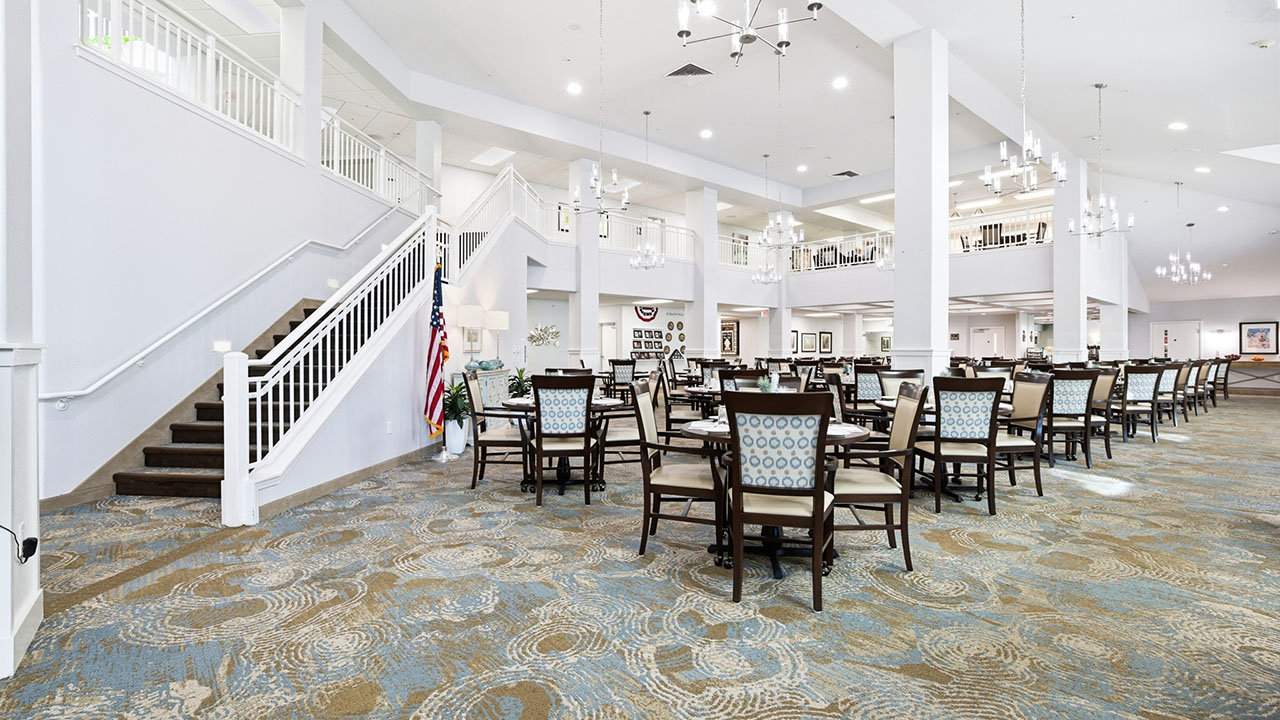 Spacious dining area with multiple tables and chairs, white columns, chandeliers, patterned carpet, and a staircase with white railings on the left side. American flags are displayed near the staircase.