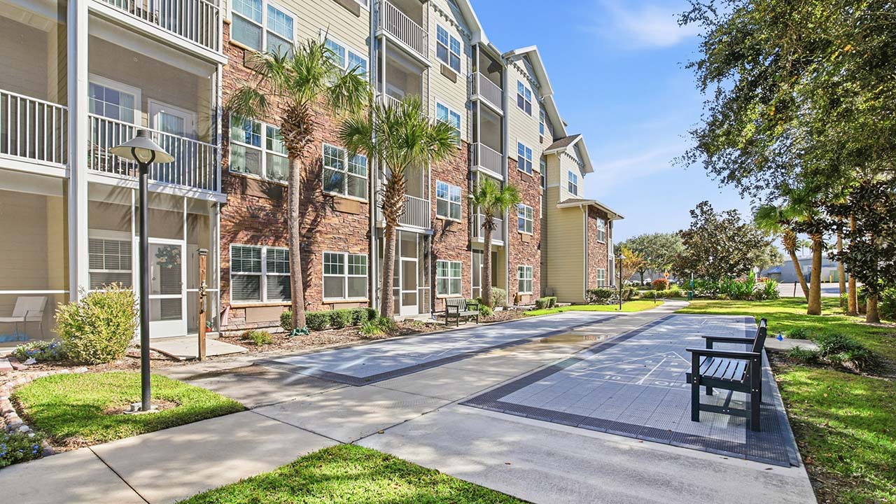 A four-story apartment building with balconies, surrounded by palm trees and greenery. In the foreground, there is a shuffleboard court and a bench along a paved walkway under a sunny blue sky.