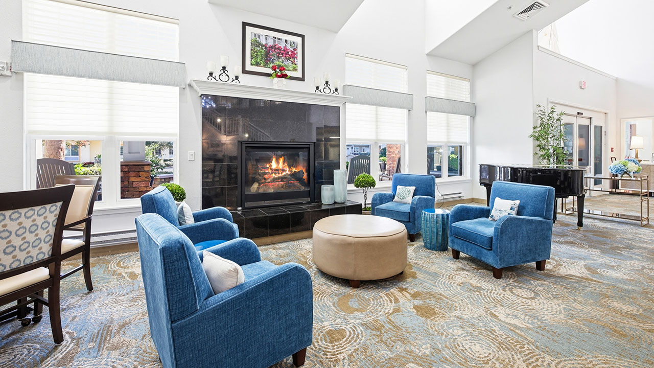 Bright, modern living room with four blue armchairs around a round ottoman, a fireplace with a fire burning, large windows, piano, and patterned carpet. Light streams in, creating a warm, inviting atmosphere.