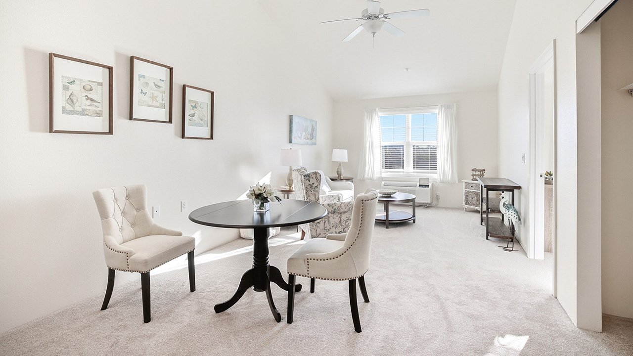 A bright, spacious living and dining area with cream walls and carpet. Two upholstered chairs sit around a small round table. Armchairs, artwork, and large windows with sheer curtains complete the airy, inviting space.