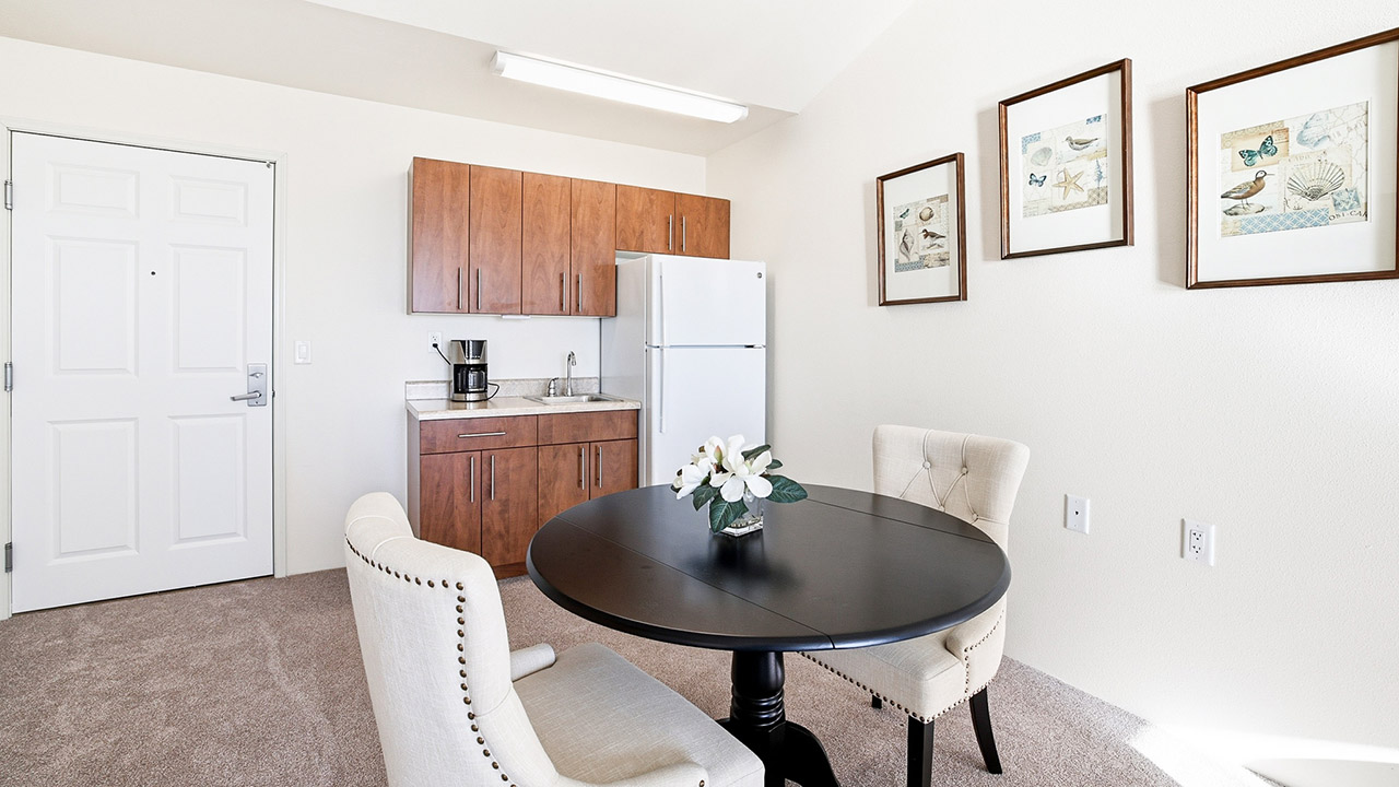 A small kitchen area with wooden cabinets, a white refrigerator, and a coffee maker. In front is a round black table with two upholstered chairs and a flower arrangement. Three framed prints hang on the wall.