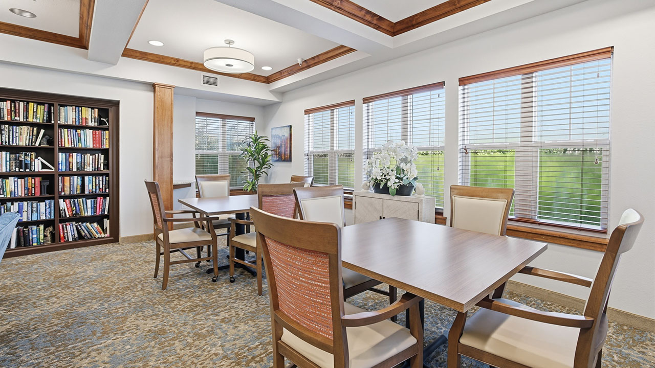 A bright room with large windows, wooden tables and chairs, a bookshelf filled with books, decorative plants, and beige carpeting. Sunlight streams in, creating a welcoming atmosphere.
