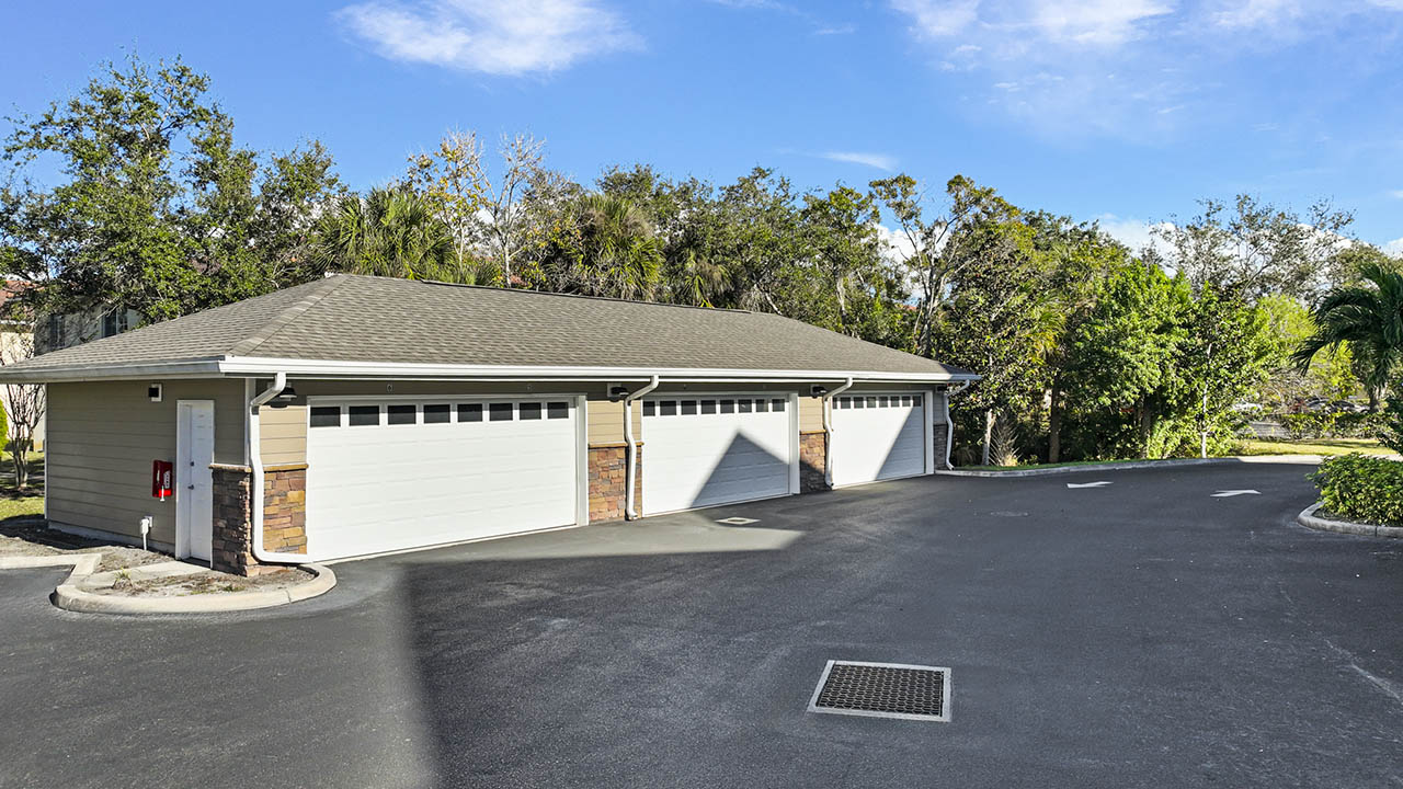 A row of four closed white garage doors in a single building, surrounded by trees and greenery, with a paved driveway and a clear blue sky above.