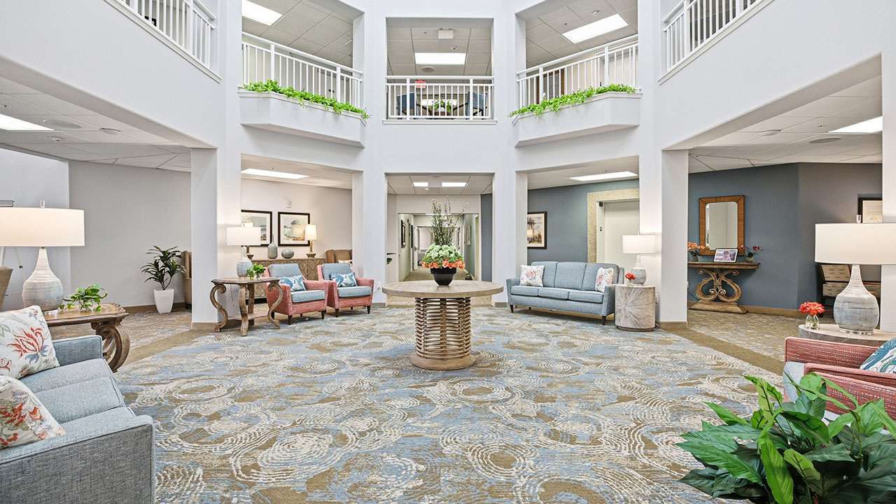 A bright, spacious lobby with modern decor features cushioned chairs and sofas, a round table with flowers in the center, potted plants, and art on light-colored walls. Skylights and balconies add openness above.