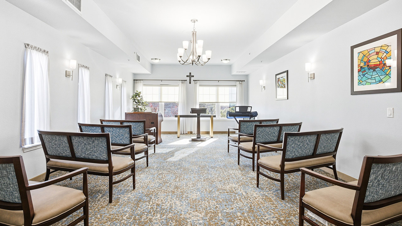 A small, bright chapel with rows of cushioned chairs facing a podium and cross. Large windows let in natural light. A keyboard, framed artwork, and a chandelier are visible in the clean, serene space.