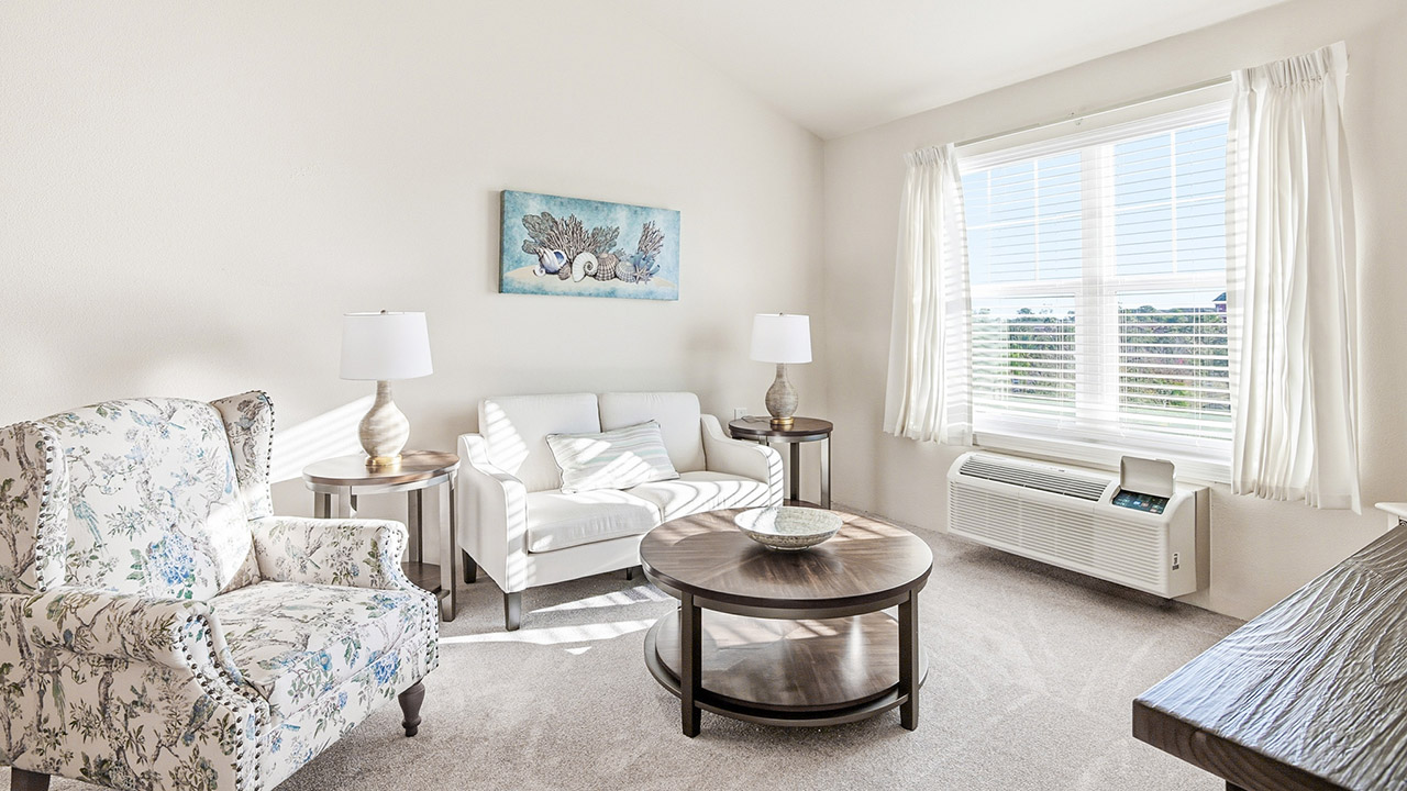 Bright living room with a floral armchair, white loveseat, two side tables with lamps, a round coffee table, large window with white curtains, wall art, and beige carpeted floor. Sunlight fills the airy space.