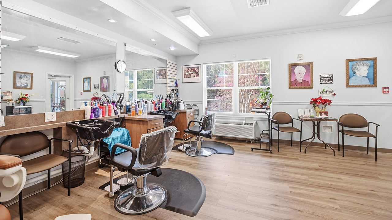 A bright, tidy hair salon with empty styling chairs, hair products on the counter, framed photos on the walls, large windows letting in natural light, and a small seating area with two chairs and a table.