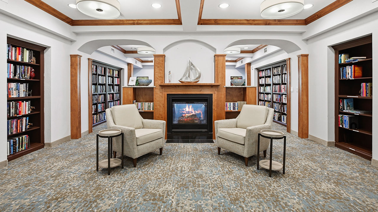 A cozy library reading area with two beige armchairs and round side tables facing a modern fireplace, surrounded by bookshelves filled with books. A decorative sailboat sits on the mantel above the fireplace.
