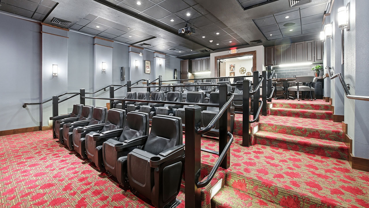 A small private movie theater with rows of black cushioned seats, carpeted red floral floor, soft wall lighting, and a large screen at the front; the space appears modern and inviting.