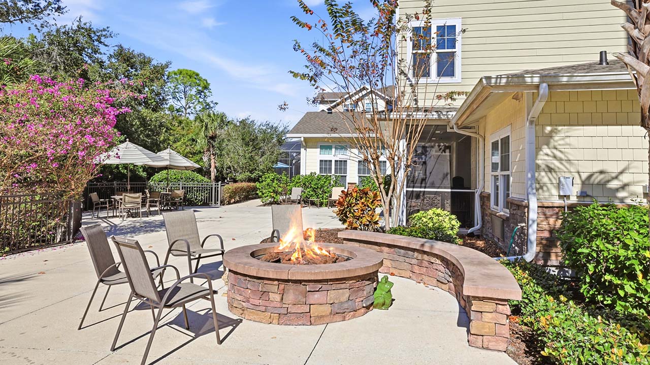 A sunny patio with metal chairs and a brick fire pit, surrounded by lush greenery and a curved stone bench, is next to a light-colored house with large windows.