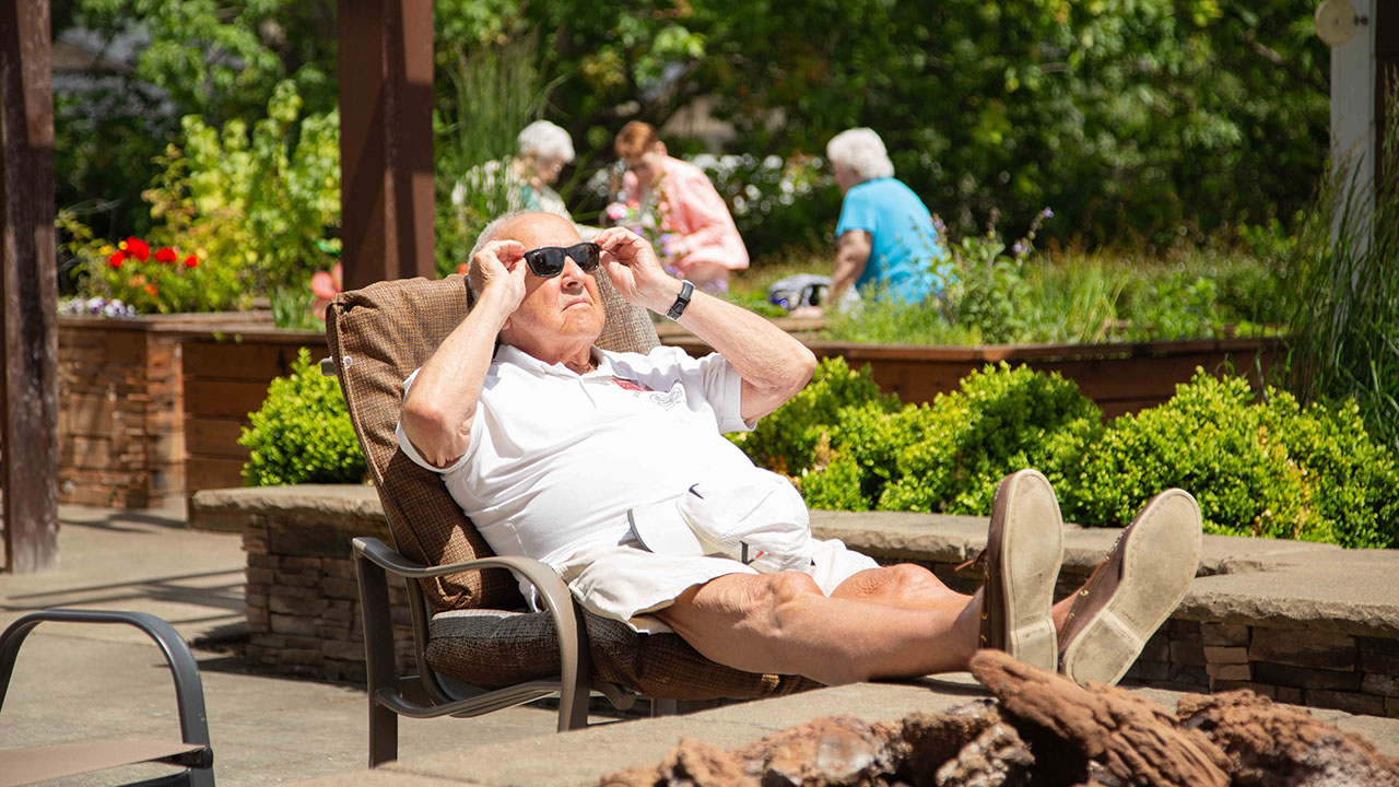 An elderly man in white clothes relaxes in an outdoor chair, adjusting his sunglasses. He is in a sunny garden area with greenery, while a few other people socialize in the background.