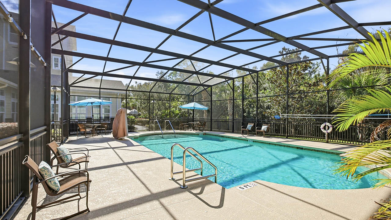 A screened-in outdoor swimming pool with metal railing, patio chairs, pool slide, and tables with blue umbrellas. Palm trees and greenery are visible outside the enclosure.