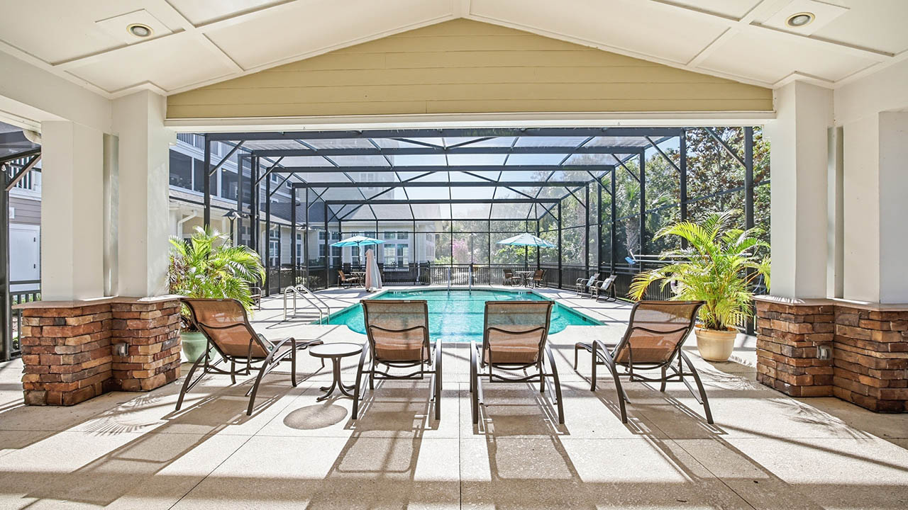 Four lounge chairs face a screened-in outdoor pool with turquoise water. Potted plants sit near the pool, and patio tables with umbrellas are in the background under a covered patio roof.