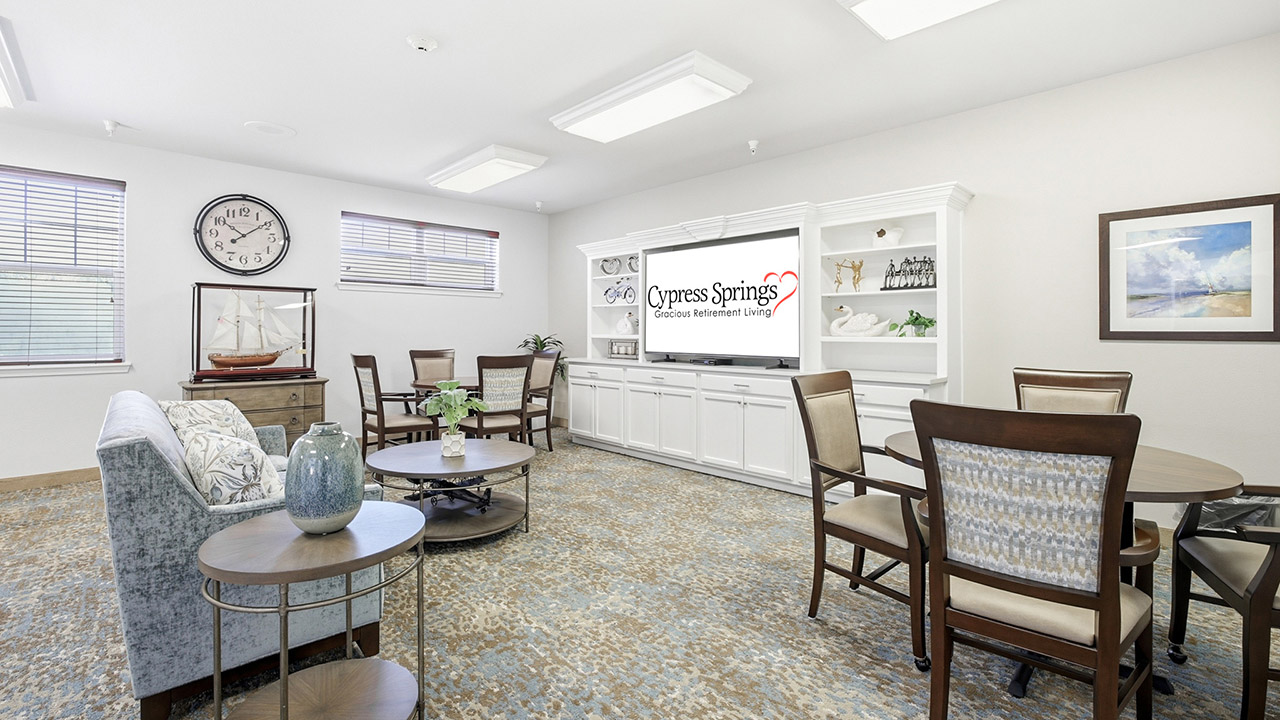 A bright, spacious lounge with armchairs, round tables, a clock, ship model, and built-in shelves. A TV displays "Cypress Springs Gracious Retirement Living." Large windows let in natural light.