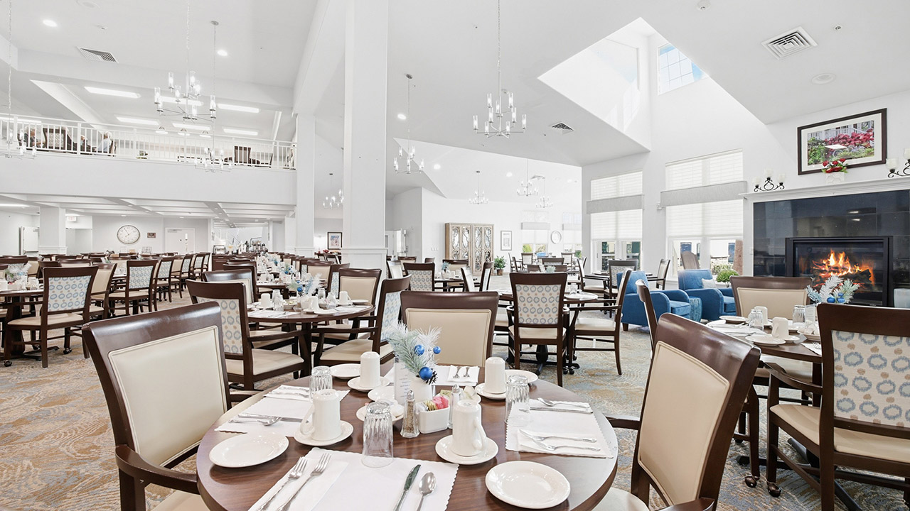 A spacious, bright dining hall with neatly set tables and cushioned chairs, featuring high ceilings, chandeliers, large windows, and a cozy fireplace; some lounge chairs are visible in the background.