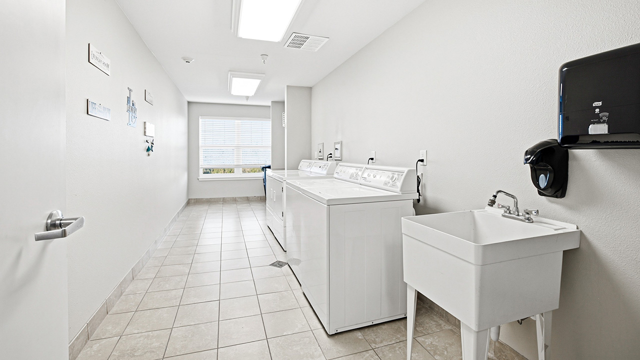 A clean, bright laundry room with two washing machines, a dryer, a utility sink, tiled floor, white walls, and a large window letting in natural light. A paper towel dispenser is mounted on the right wall.