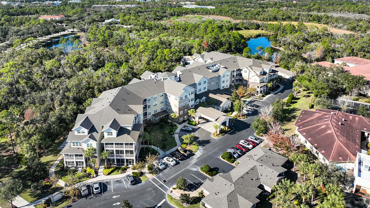 Aerial view of a large residential complex surrounded by trees and greenery, with parking lots and multiple parked cars. A pond and additional buildings are visible in the background.
