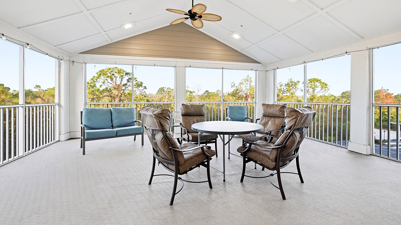 A spacious screened-in porch with a round table, five cushioned chairs, a blue loveseat, ceiling fan, and railing, overlooking trees and greenery outside.