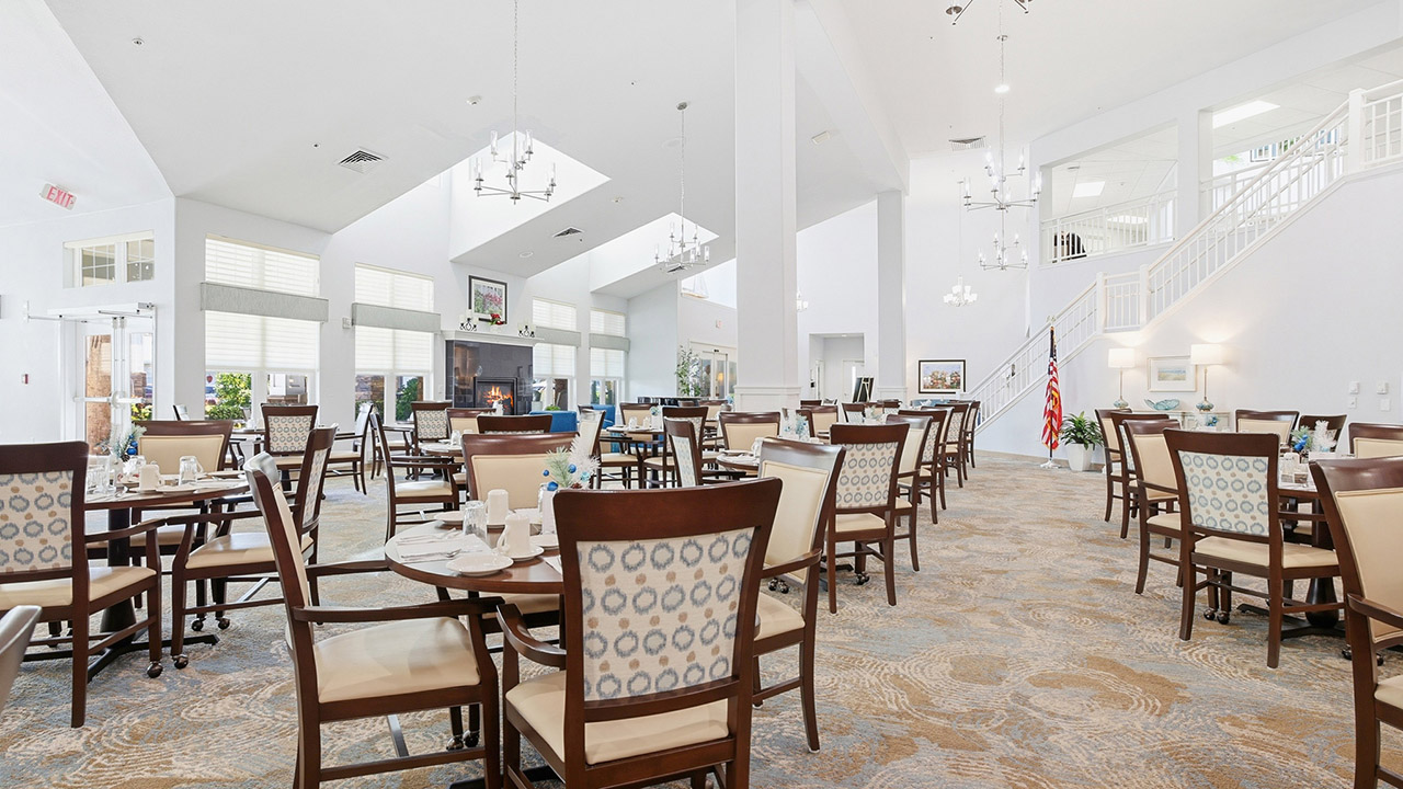 Bright, spacious dining room with many neatly set tables and cushioned chairs, large windows letting in natural light, chandeliers hanging from the ceiling, and an American flag near a white staircase.