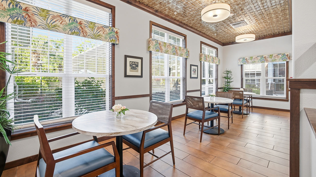 A bright café-style room with wooden floor, round marble tables, wooden chairs with dark cushions, large windows with floral valances, and decorative wall art. Sunlight streams through the windows, creating a welcoming atmosphere.