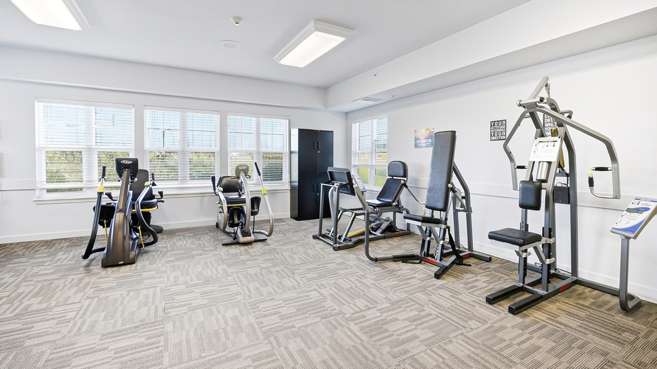 A bright, modern gym room with four exercise machines, light-colored carpet, large windows with blinds, and a black cabinet against the back wall. The room is well-lit and neatly organized.