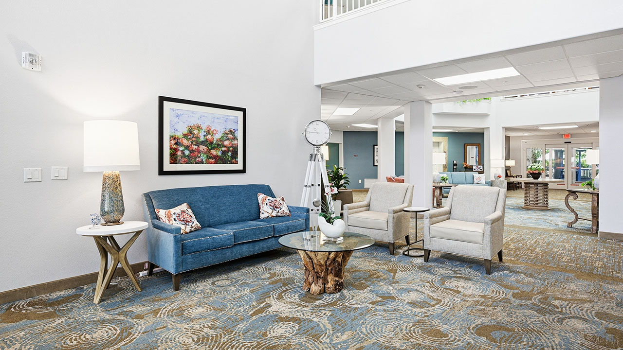 A bright, modern lounge area with a blue sofa, two beige armchairs, a glass coffee table with a wooden base, a lamp, wall art, a large clock, and patterned carpet in an open, airy space.