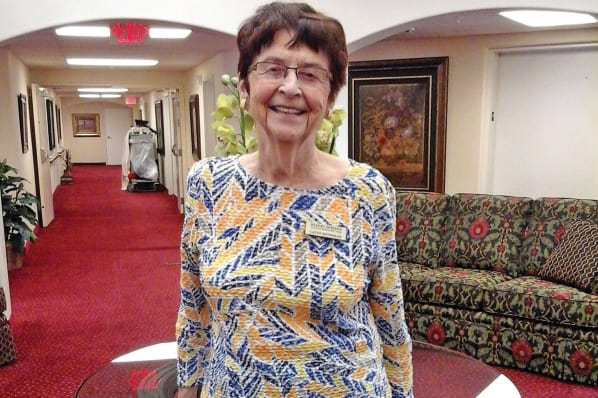 An elderly woman with short dark hair and glasses is standing in a hallway with red carpet and a patterned couch. She is smiling and wearing a yellow, blue, and white patterned top with a name tag. The hallway features plants, artwork, and arched doorways.