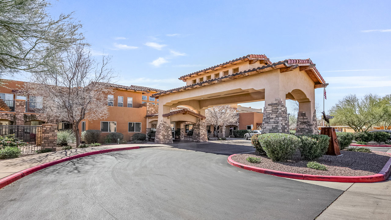 A large, tan stucco building with a covered driveway entrance, stone pillars, red tile roof, and surrounding desert landscaping under a clear blue sky.