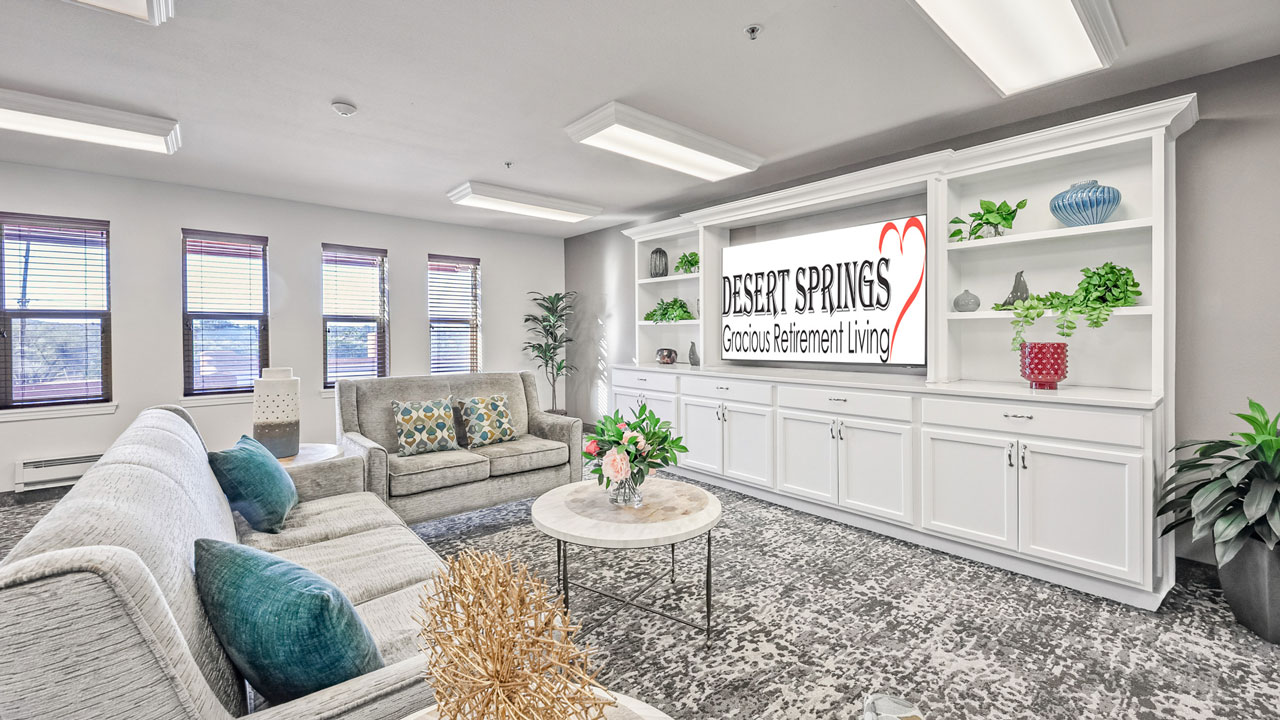 A bright, modern living room with white shelves, a large TV displaying "Desert Springs Gracious Retirement Living," gray sofas with teal and patterned cushions, a round coffee table, and large windows with blinds.