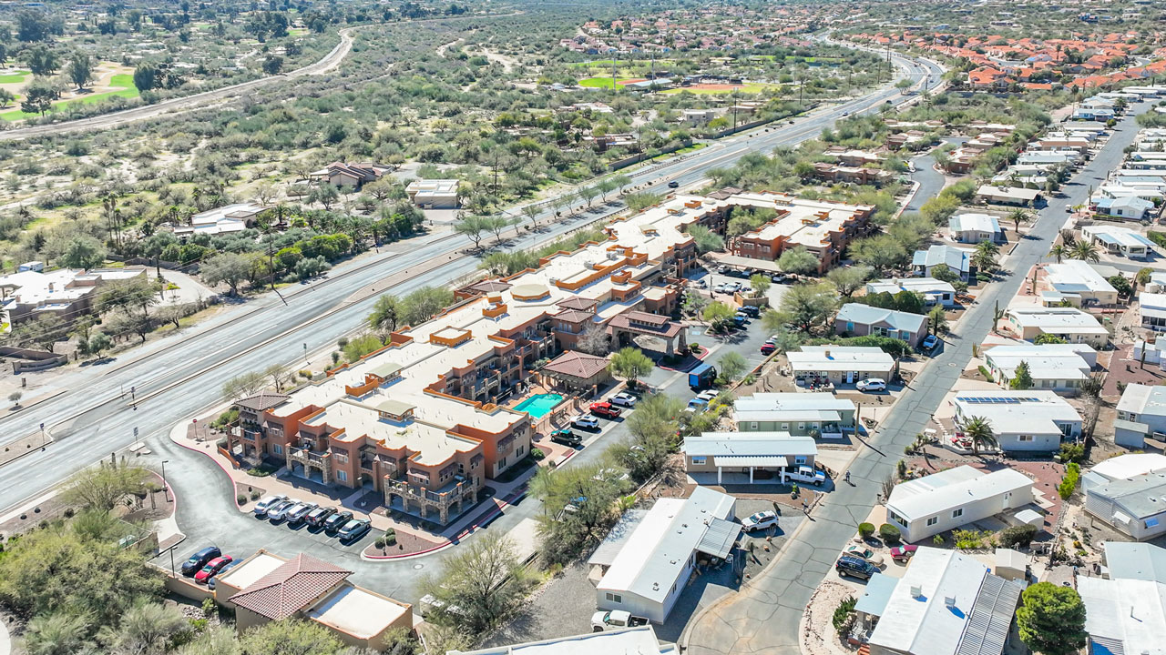 Aerial view of a desert community with a large southwestern-style apartment complex, a swimming pool, parking lots, mobile homes, and a nearby four-lane road surrounded by sparse vegetation and distant residential areas.