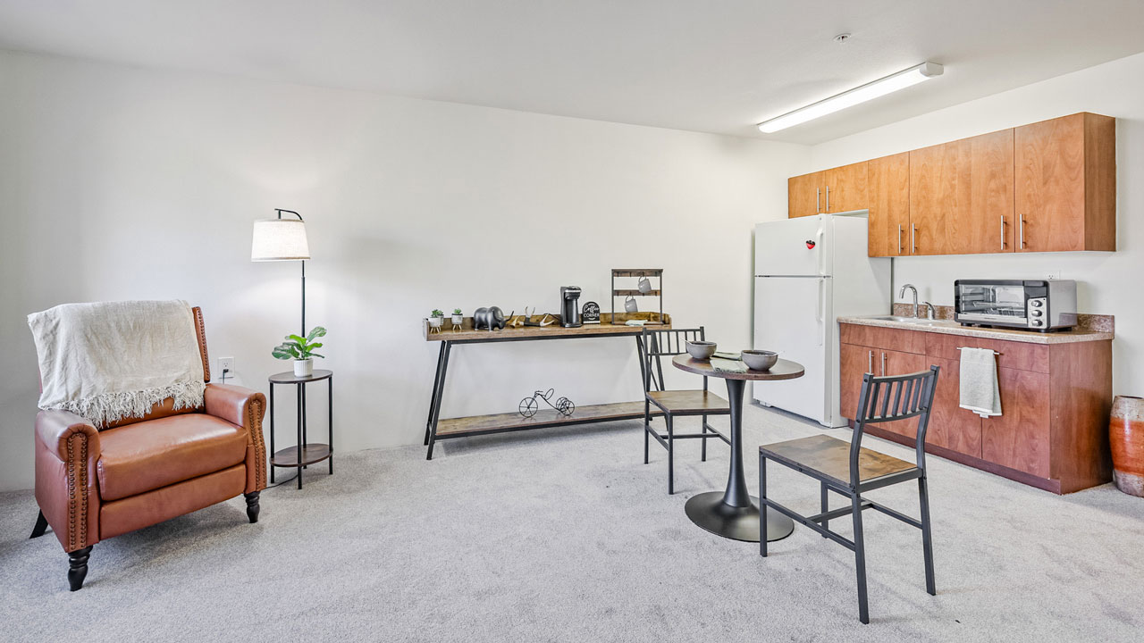 A cozy studio with a brown leather armchair, round dining table with two chairs, a console table with decor, and a compact kitchen with wood cabinets, white appliances, and a microwave on the counter.